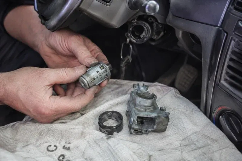 Hands holding a removed ignition cylinder beside a car steering column, with the housing and small parts on a cloth. — Ignition Lock Repair, Niagara Falls