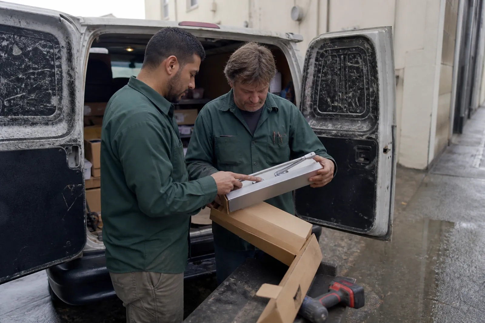 Two technicians in green shirts look over a metal tray holding a chrome tool beside a white van with open rear doors. — Royal